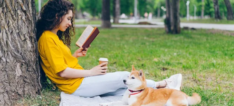 Person reading a book and holding a coffee by a tree with a Shiba Inu dog beside them in a park